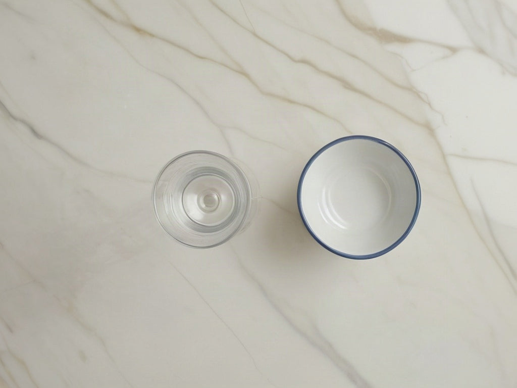 Overhead view of a tall glass of water and a small white ceramic dish on a light marble countertop