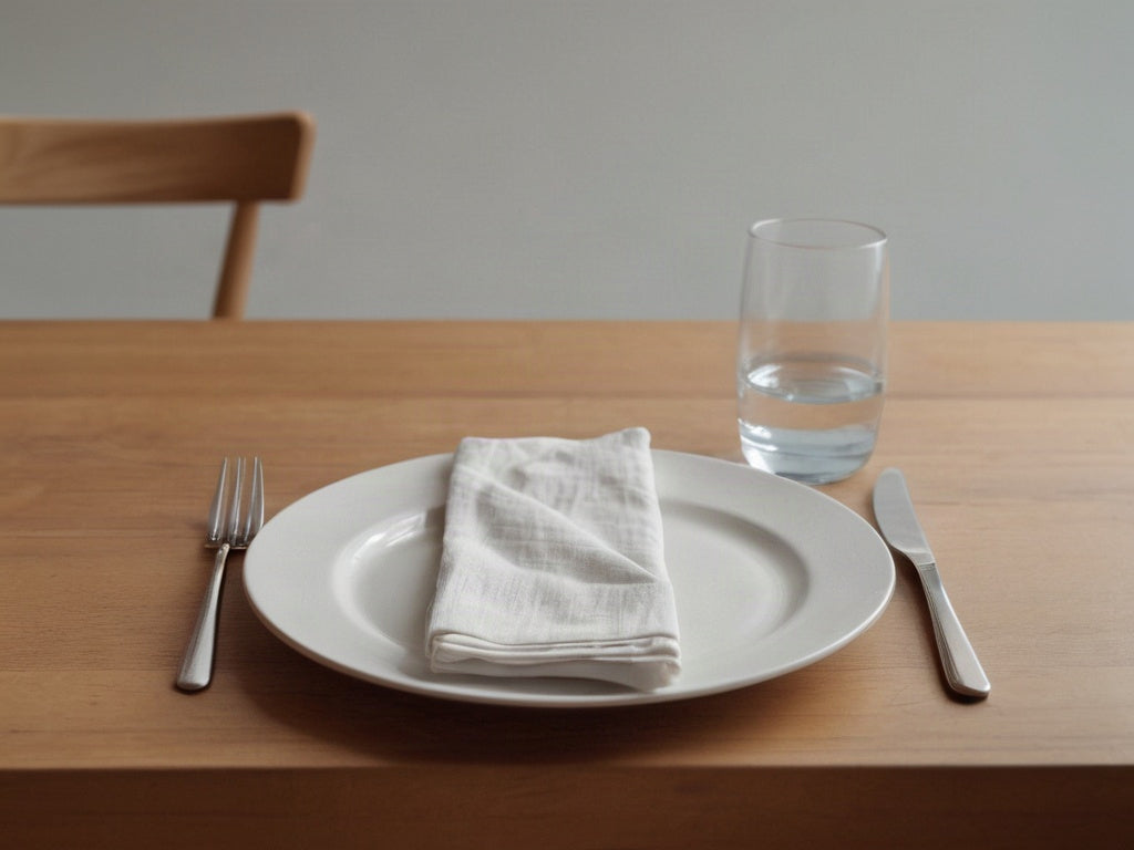 An empty white plate with a folded linen napkin, fork, knife, and a glass of water on a wooden dining table