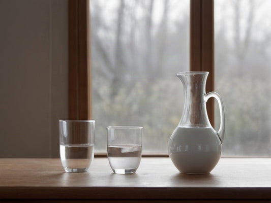 A white ceramic pitcher and two glasses of water on a wooden table beside a window in soft morning light
