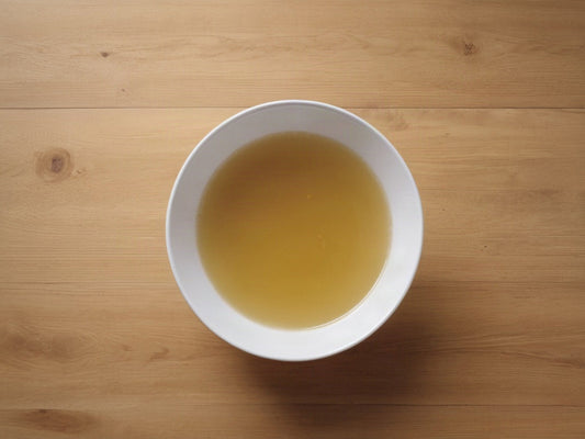 Overhead shot of clear golden broth in a plain white bowl on a wooden table